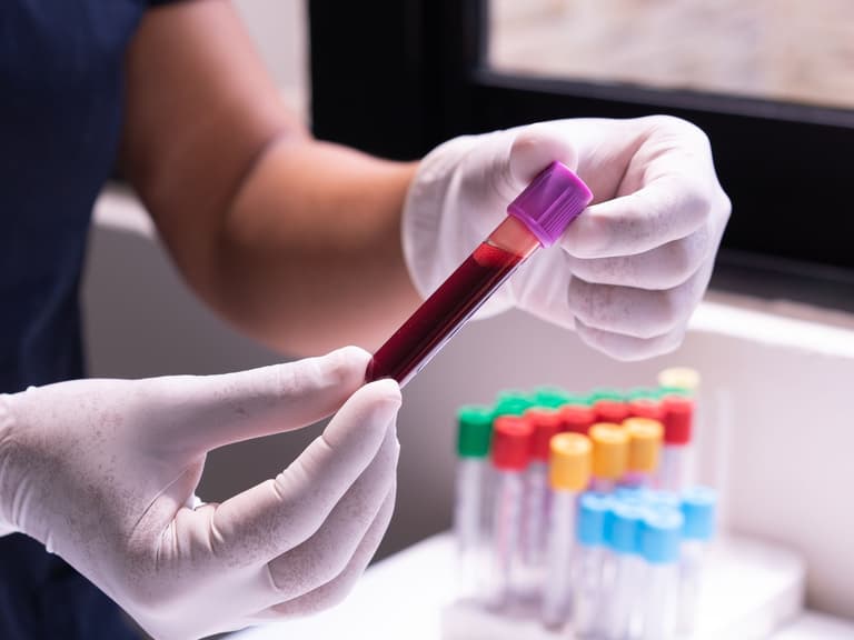 Mains gantées tenant un tube de prélèvement sanguin au-dessus d'un portoir de tubes